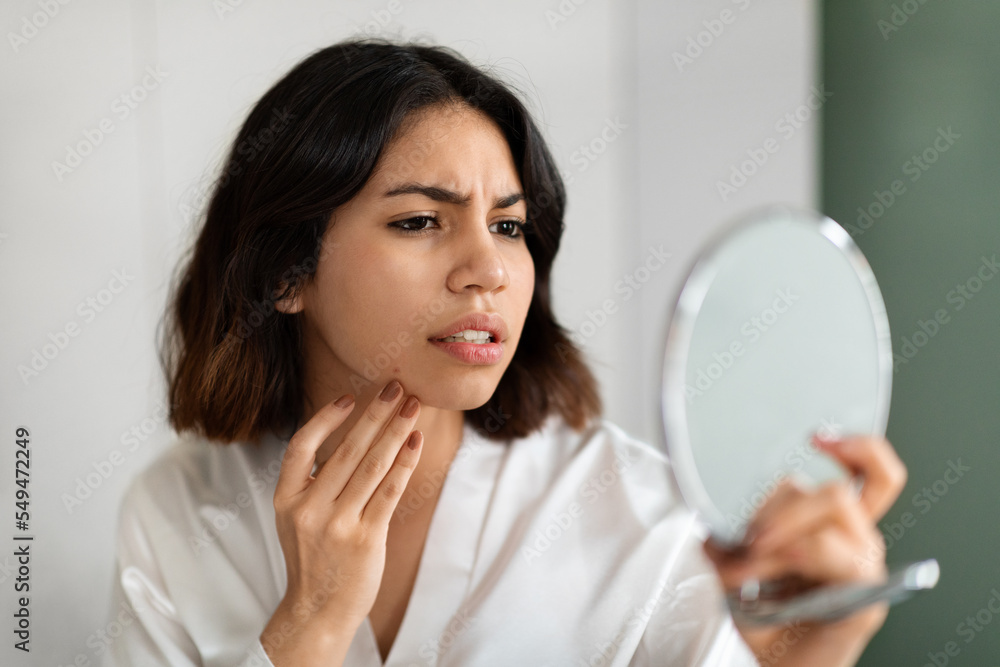 Upset young woman looking at mirror and touching her face Upset young woman looking at mirror and touching her face