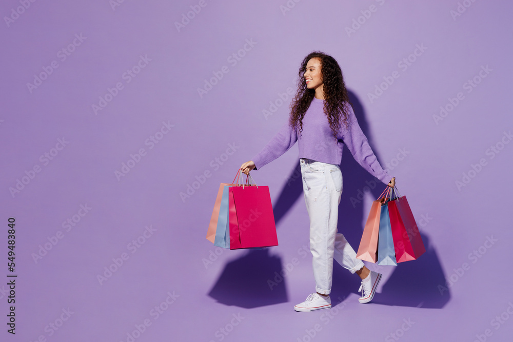 Full body side profile view young woman wear pullover hold in hand paper package bags after shopping walking going isolated on plain pastel light purple background. Black Friday sale buy day concept. Full body side profile view young woman wear pullover hold in hand paper package bags after shopping walking going isolated on plain pastel light purple background. Black Friday sale buy day concept.