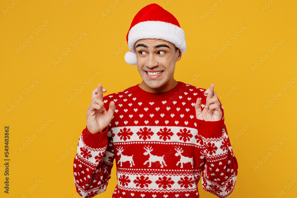 Merry young man wear red knitted christmas sweater Santa hat posing wait special moment keeping fingers crossed, making wish hope trust isolated on plain yellow background Happy New Year 2023 concept Merry young man wear red knitted christmas sweater Santa hat posing wait special moment keeping fingers crossed, making wish hope trust isolated on plain yellow background Happy New Year 2023 concept