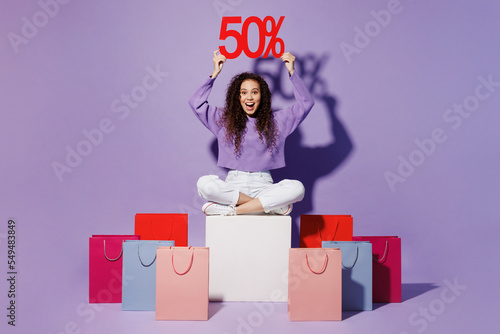 Full body young woman wear pullover sit near shopping paper package bag afer hold overhead 50 percent discount sign isolated on plain pastel light purple background Black Friday sale buy day concept. photo