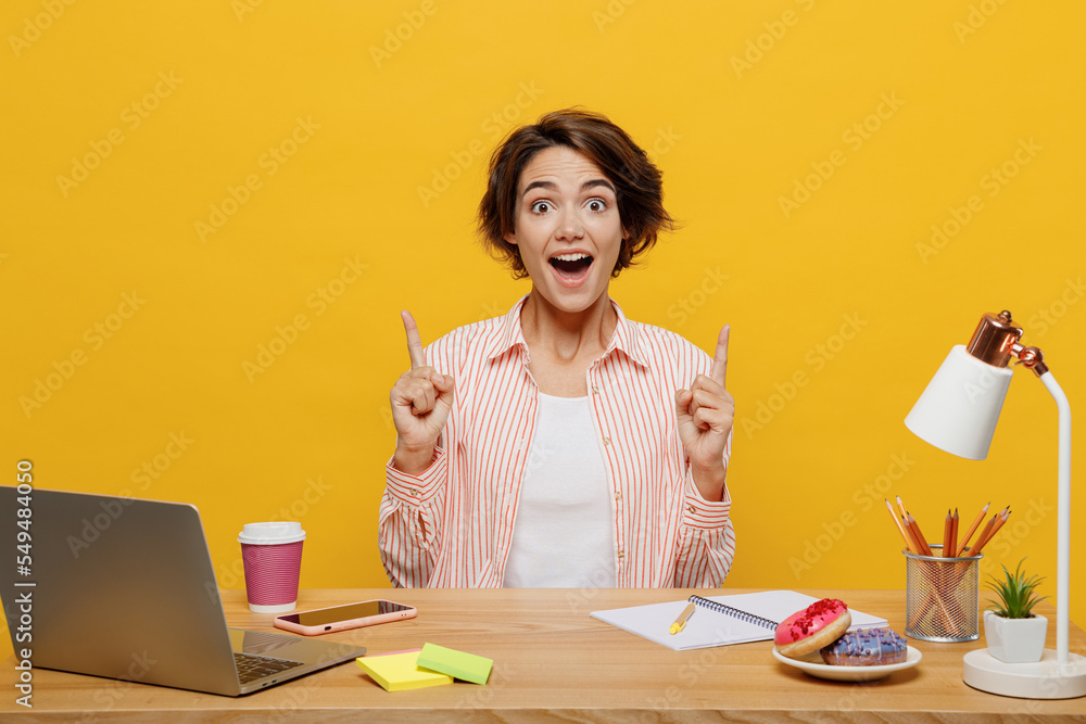 Young surprised employee business woman wear casual shirt sit work at office desk with pc laptop point fingers overhead on area isolated on plain yellow color background. Achievement career concept.