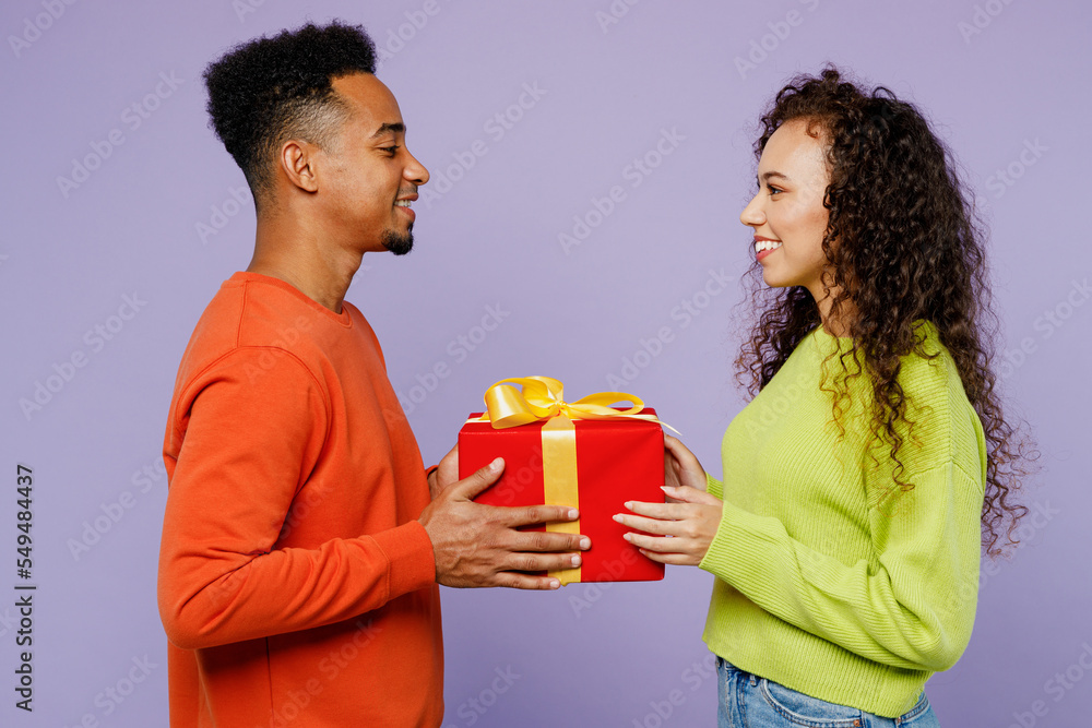 Side view young couple two friend family man woman of African American ethnicity in casual clothes together give red present box with gift ribbon bow isolated on pastel plain light purple background Side view young couple two friend family man woman of African American ethnicity in casual clothes together give red present box with gift ribbon bow isolated on pastel plain light purple background