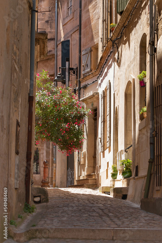 A typical, narrow, picturesque street in the Provence region of France. A street with building facades and colorful flowers in the city of Arles. Summer in the Mediterranean region. © inguunal