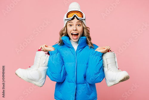 Snowboarder surprised happy woman wear blue suit goggles mask hat ski padded jacket hold boots in hand isolated on plain pastel pink background Winter extreme sport hobby weekend trip relax concept.