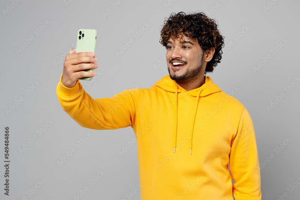 Young smiling Indian man 20s wear casual yellow hoody doing selfie shot on mobile cell phone post photo on social network isolated on plain grey background studio portrait. People lifestyle portrait. Young smiling Indian man 20s wear casual yellow hoody doing selfie shot on mobile cell phone post photo on social network isolated on plain grey background studio portrait. People lifestyle portrait.