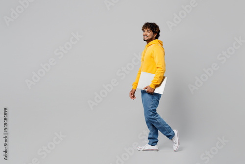 Side profile full body view young happy smiling fun IT Indian man 20s wear casual yellow hoody hold closed laptop pc computer walking going strolling isolated on plain grey background studio portrait.