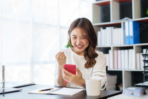 Beautiful Asian businesswoman using smartphone is delighted and excited by the success she has achieved through her office cell phone.