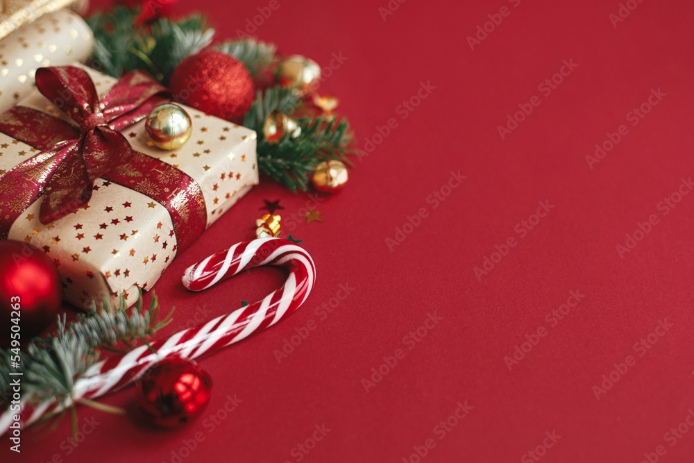 Modern christmas composition. Stylish christmas gift, wrapping paper,  fir branches, candy cane and baubles on red background, space for text. Merry Christmas and Happy Holidays! Festive banner