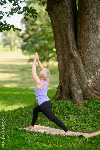 Middle aged woman doing yoga outdoors in warrior pose, Virabhadrasana I. Sports concept in city park. © GRON777