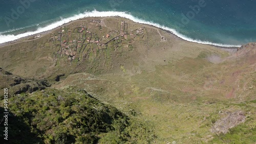 Aerial bird's eye view of an amazing cliff next to the ocean. Small village between the sea and a huge cliff. Flying forward. Located in Madeira, Portugal.