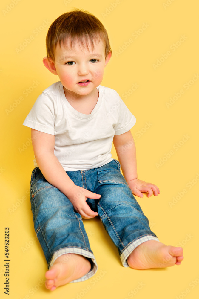 Portrait of an embarrassed toddler baby in full length on a studio yellow background. The child is sitting on the floor in a white T-shirt and blue jeans. Kid aged one year and four months Portrait of an embarrassed toddler baby in full length on a studio yellow background. The child is sitting on the floor in a white T-shirt and blue jeans. Kid aged one year and four months