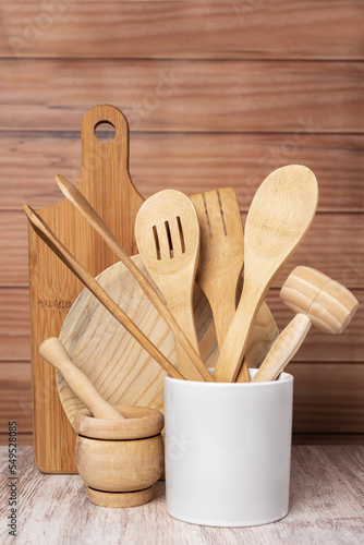 Wooden kitchen utensils, plate, cutting board and mortar, next to a jar with a spoon, fork and tongs among others, decoratively arranged with a light wooden background. Copy space.