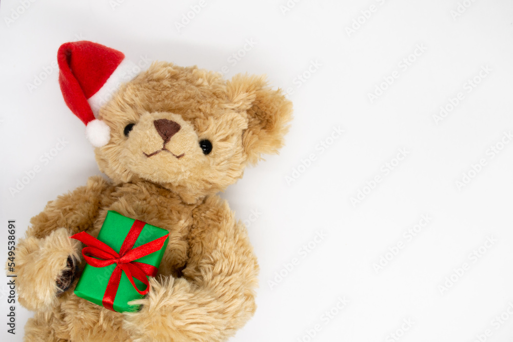 A stuffed toy Teddy bear in a red Santa Claus hat with a pompom on one ear, holding green gift boxes in its paws. White background, copy space.