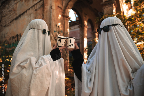 A funny image of two people in ghost costumes and sunglasses holding a cup with a drink in an abandoned building