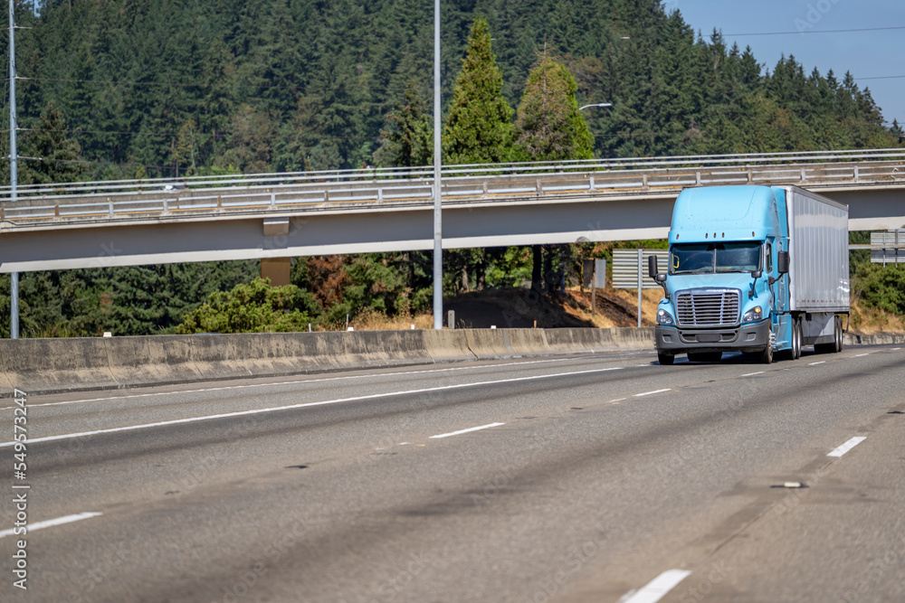 Light blue big rig semi truck with dry van semi trailer carry cargo driving on the wide highway road under the bridge