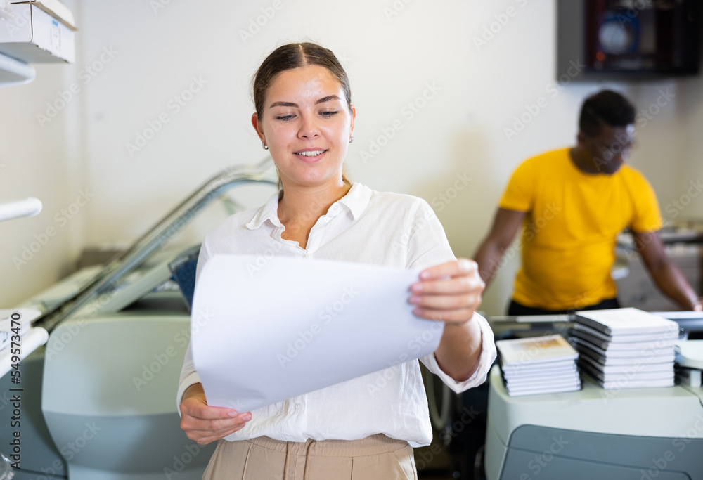 Fototapeta premium Attentive young female specialist verifying printed sheet during work in the print shop