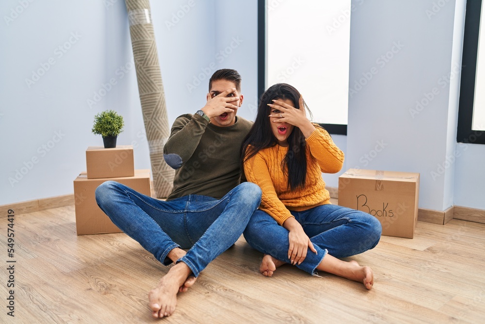 Young couple sitting on the floor at new home peeking in shock covering face and eyes with hand, looking through fingers with embarrassed expression. Young couple sitting on the floor at new home peeking in shock covering face and eyes with hand, looking through fingers with embarrassed expression.