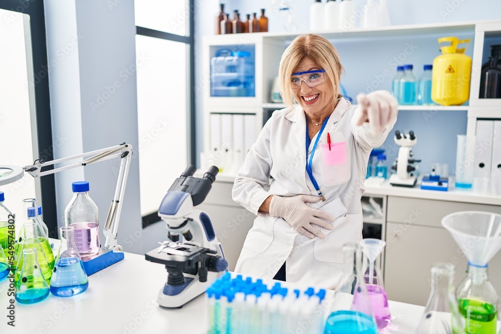 Middle age blonde woman working at scientist laboratory laughing at you, pointing finger to the camera with hand over body, shame expression Middle age blonde woman working at scientist laboratory laughing at you, pointing finger to the camera with hand over body, shame expression