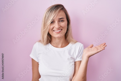 Young blonde woman standing over pink background smiling cheerful presenting and pointing with palm of hand looking at the camera.