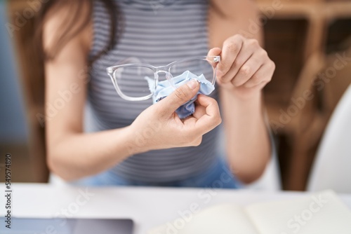 Young hispanic girl cleaning glasses sitting on table at home