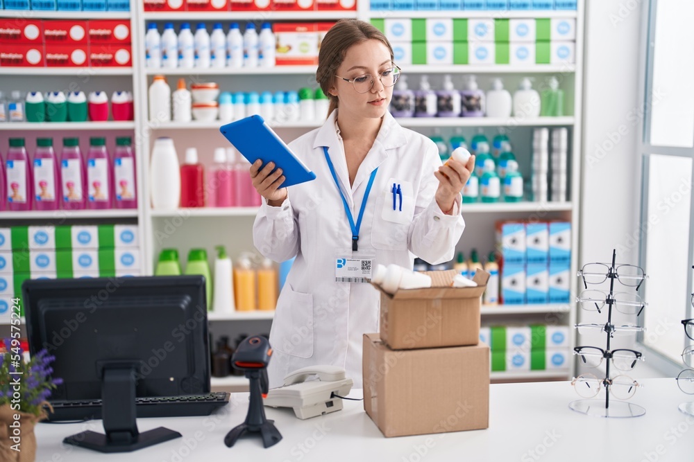 Young blonde woman pharmacist using touchpad holding pills bottle at pharmacy