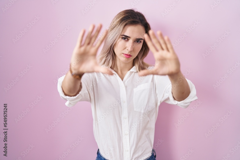Young beautiful woman standing over pink background doing frame using hands palms and fingers, camera perspective Young beautiful woman standing over pink background doing frame using hands palms and fingers, camera perspective