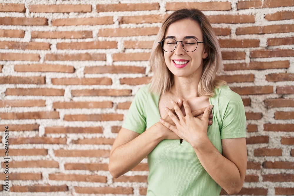 Young beautiful woman standing over bricks wall smiling with hands on chest with closed eyes and grateful gesture on face. health concept. Young beautiful woman standing over bricks wall smiling with hands on chest with closed eyes and grateful gesture on face. health concept.