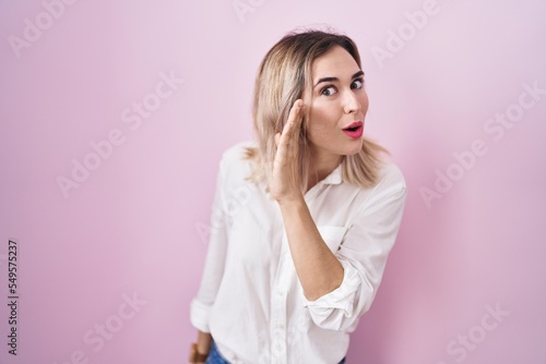 Young beautiful woman standing over pink background hand on mouth telling secret rumor, whispering malicious talk conversation