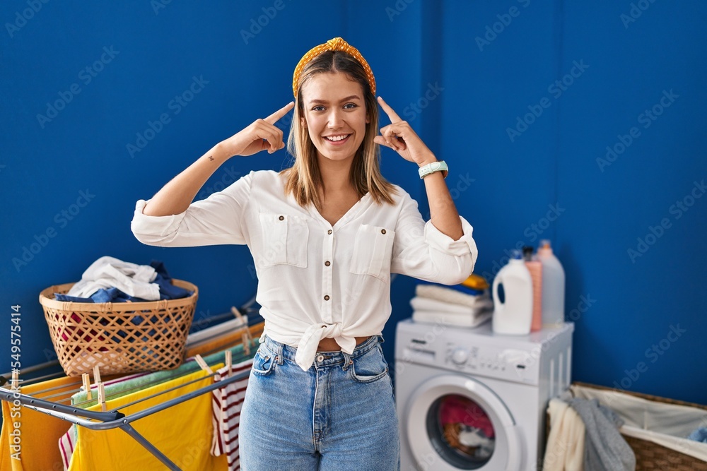 Young blonde woman at laundry room smiling pointing to head with both hands finger, great idea or thought, good memory