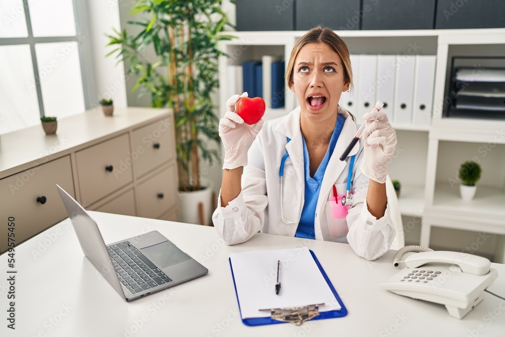 Young blonde woman wearing doctor uniform holding blood and heart angry and mad screaming frustrated and furious, shouting with anger looking up. Young blonde woman wearing doctor uniform holding blood and heart angry and mad screaming frustrated and furious, shouting with anger looking up.