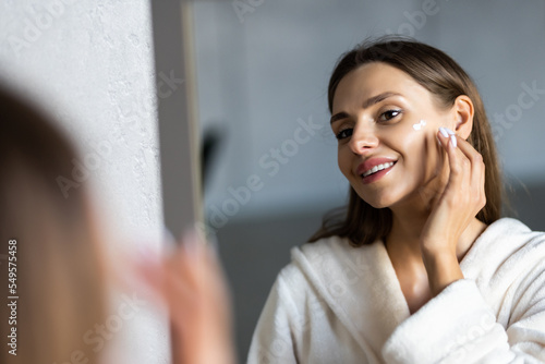 Close up cropped portrait of a young beautiful woman in bath towel smiling applying face body cream on her face for rejuvenation soft moisturizing effect. Spa beauty treatment concept. Body skin care