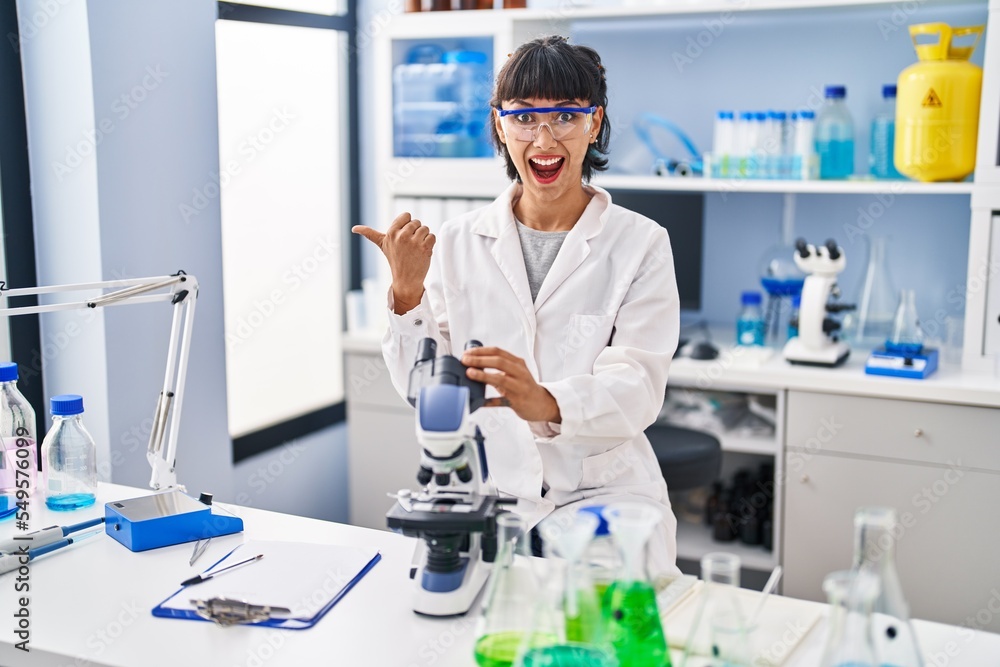 Young hispanic woman working at scientist laboratory pointing thumb up to the side smiling happy with open mouth Young hispanic woman working at scientist laboratory pointing thumb up to the side smiling happy with open mouth