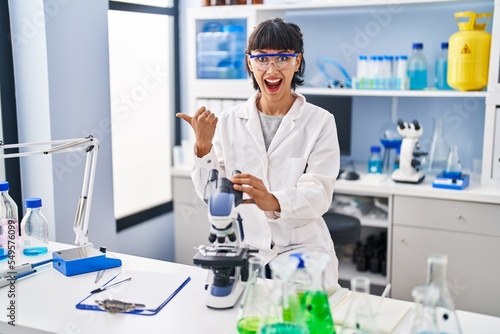 Young hispanic woman working at scientist laboratory pointing thumb up to the side smiling happy with open mouth