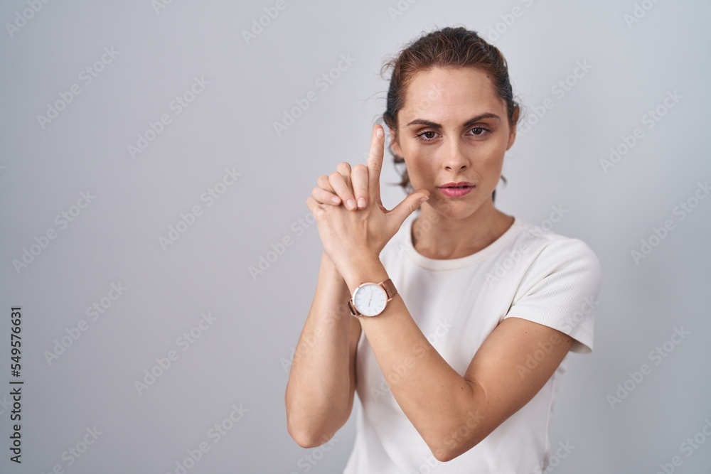 Beautiful brunette woman standing over isolated background holding symbolic gun with hand gesture, playing killing shooting weapons, angry face Beautiful brunette woman standing over isolated background holding symbolic gun with hand gesture, playing killing shooting weapons, angry face