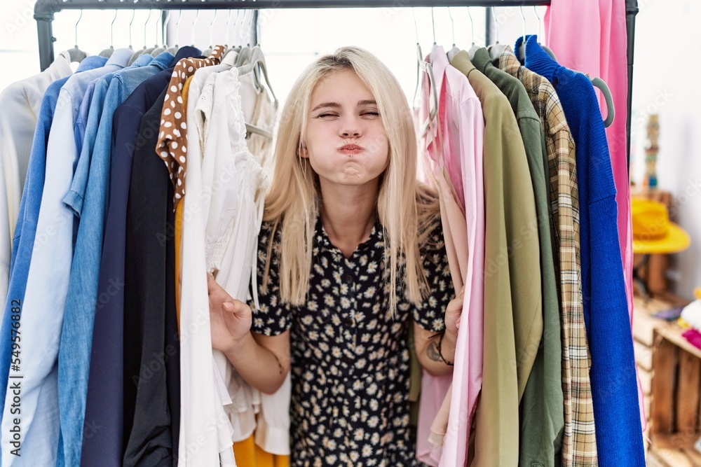 Young blonde woman searching clothes on clothing rack puffing cheeks with funny face. mouth inflated with air, catching air. - obrazy, fototapety, plakaty 