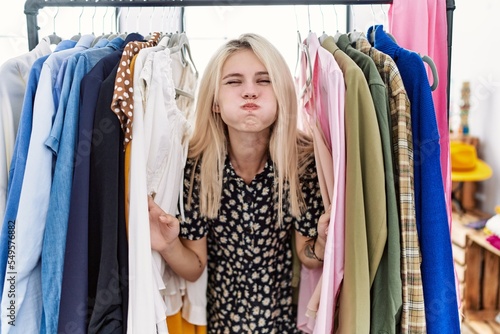Young blonde woman searching clothes on clothing rack puffing cheeks with funny face. mouth inflated with air, catching air.