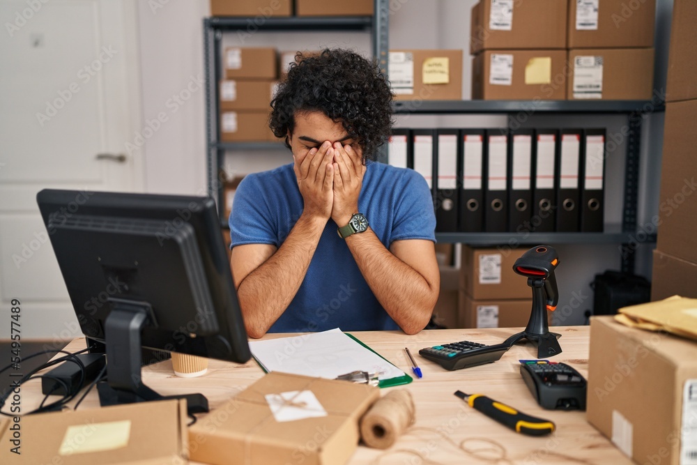 Hispanic man with curly hair working at small business ecommerce with sad expression covering face with hands while crying. depression concept.