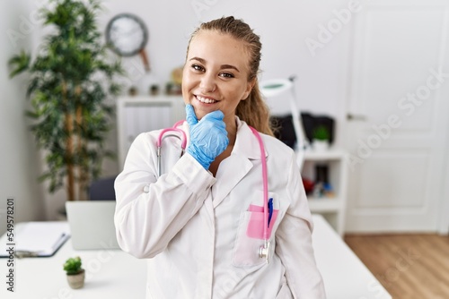 Young caucasian woman wearing doctor uniform and stethoscope at the clinic looking confident at the camera smiling with crossed arms and hand raised on chin. thinking positive.