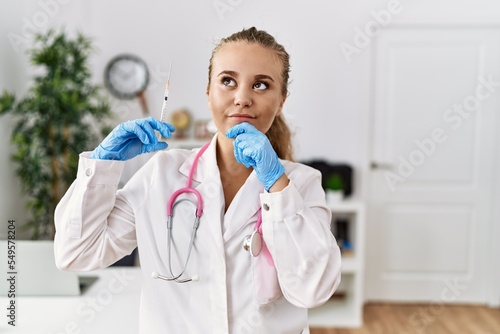 Young doctor woman holding syringe at the clinic serious face thinking about question with hand on chin  thoughtful about confusing idea