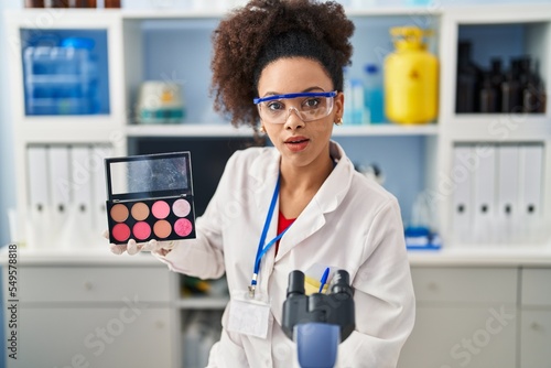 Young african american woman working at scientist laboratory doing make up scared and amazed with open mouth for surprise  disbelief face