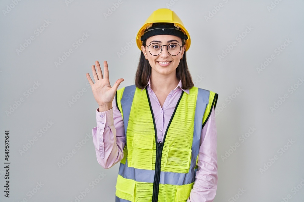 Hispanic girl wearing builder uniform and hardhat showing and pointing up with fingers number five while smiling confident and happy. Hispanic girl wearing builder uniform and hardhat showing and pointing up with fingers number five while smiling confident and happy.