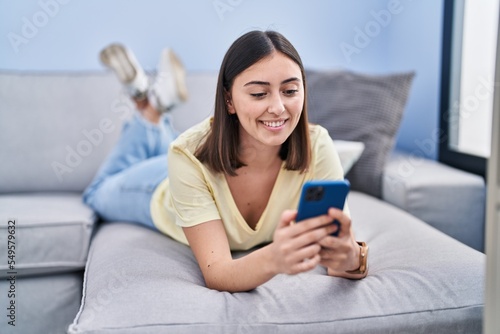 Young hispanic woman using smartphone lying on sofa at home