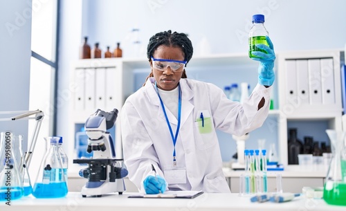 African american woman scientist holding bottle writing on document at laboratory