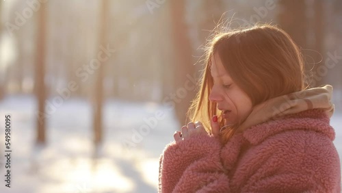 Beautiful young woman trying to warm her frozen hands with a breath in winter park. Girl with frosty breathing enjoying cold sunny weather outdoors on an icy snowy day