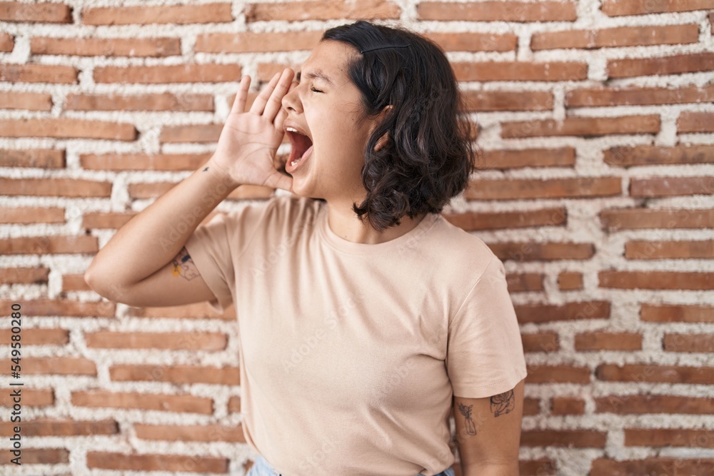 Young hispanic woman standing over bricks wall shouting and screaming loud to side with hand on mouth. communication concept.