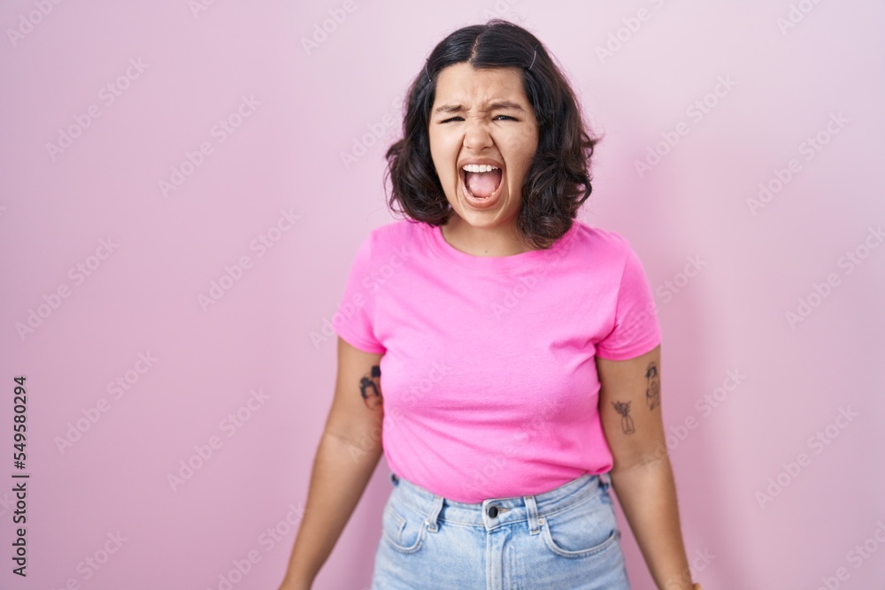 Young hispanic woman standing over pink background angry and mad screaming frustrated and furious, shouting with anger. rage and aggressive concept. Young hispanic woman standing over pink background angry and mad screaming frustrated and furious, shouting with anger. rage and aggressive concept.