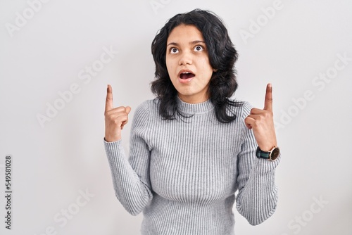Hispanic woman with dark hair standing over isolated background amazed and surprised looking up and pointing with fingers and raised arms.