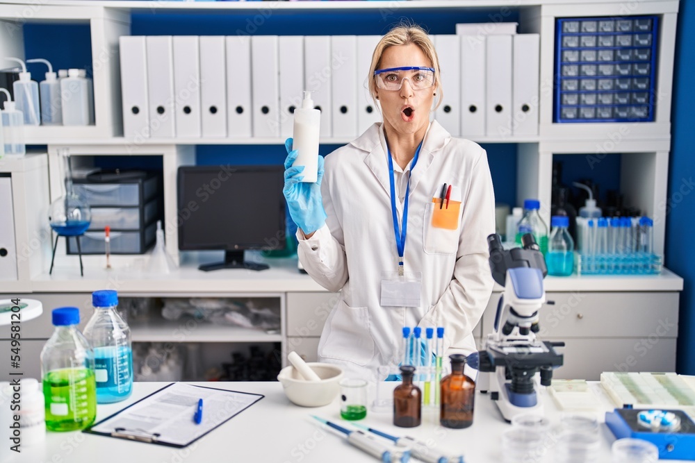 Young caucasian woman working at scientist laboratory holding body lotion scared and amazed with open mouth for surprise, disbelief face Young caucasian woman working at scientist laboratory holding body lotion scared and amazed with open mouth for surprise, disbelief face