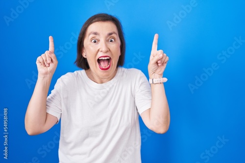 Middle age hispanic woman standing over blue background smiling amazed and surprised and pointing up with fingers and raised arms.