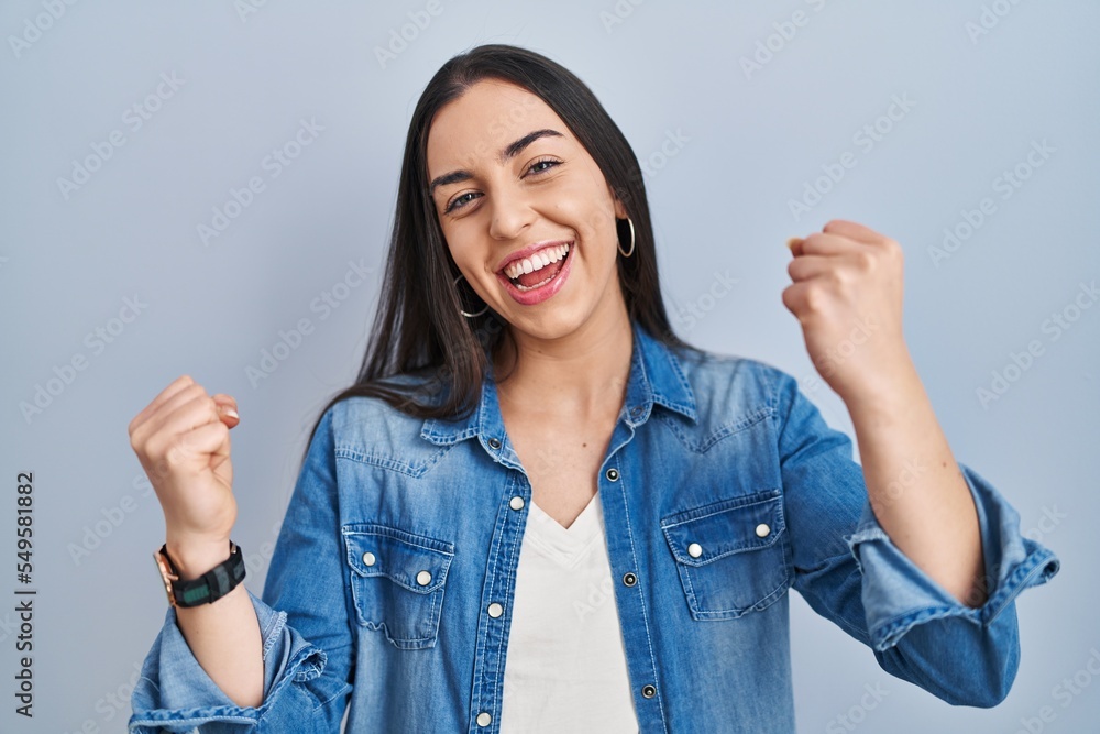 Hispanic woman standing over blue background very happy and excited doing winner gesture with arms raised, smiling and screaming for success. celebration concept. Hispanic woman standing over blue background very happy and excited doing winner gesture with arms raised, smiling and screaming for success. celebration concept.
